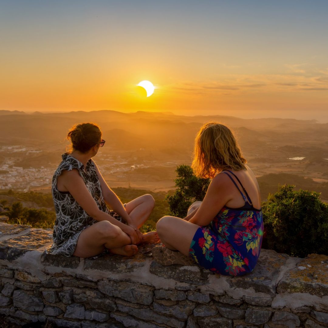 Two young female friends in summer dresses sitting on a stone wall watching a spectacular sunset during a solar eclipse from the Monte Toro viewpoint in Menorca, Spain